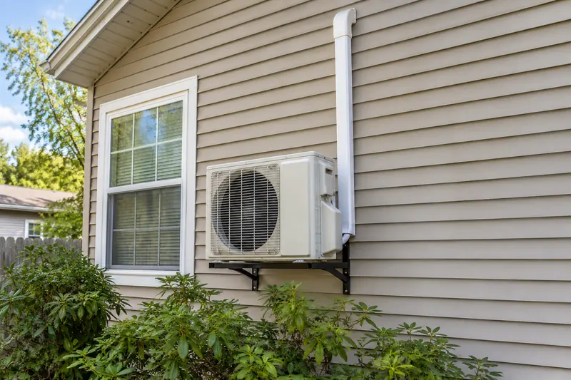 A small ductless outdoor unit on a wall bracket beside a window, its insulated line set climbing the siding