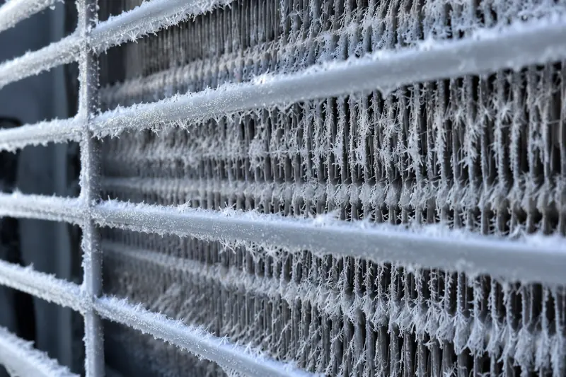 Frosted aluminum fins on a heat pump's outdoor coil seen up close, the white crust thin enough to show the metal beneath
