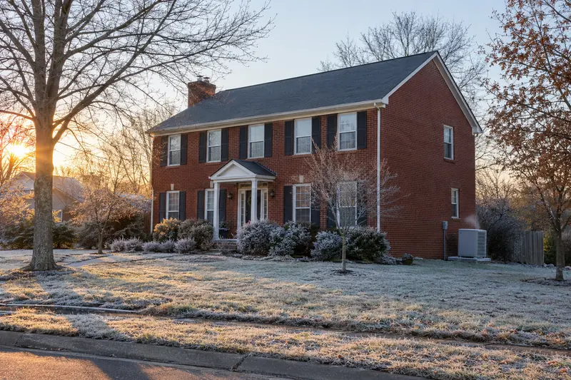 Two-story red-brick house on a frosty sunrise lawn under bare trees, a heat pump's outdoor unit at its side