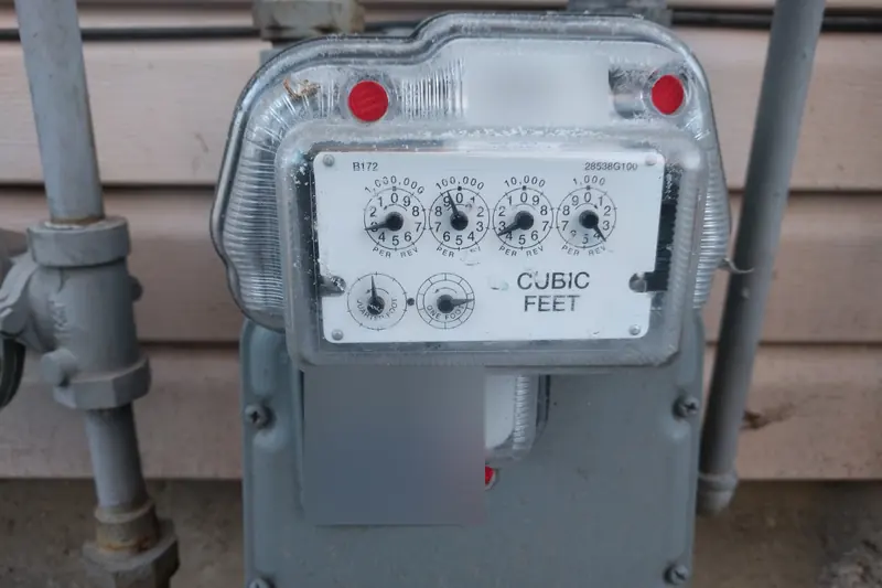 A natural gas meter with its gray regulator and piping against the outside wall of a house