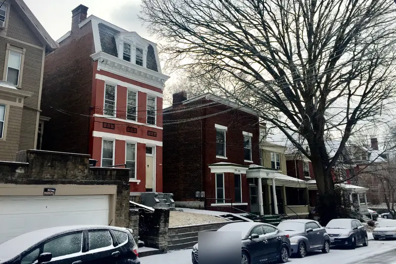 A city street of brick row houses after a snowfall, snow on parked cars and a large bare tree